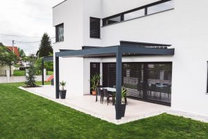 Modern house exterior with a clean, minimalist design featuring smooth acrylic render walls, large windows with black shutters, and a sleek outdoor patio with a pergola. The patio includes a dining table, chairs, and potted plants, surrounded by a well-manicured lawn.