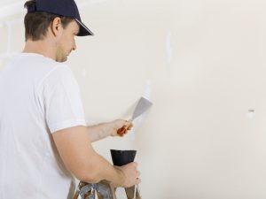 Person patching wall cracks with a scraper, applying putty to fix surface damage in a white room.